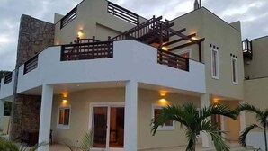 A modern two-story house with a stone facade, a balcony with a wooden railing, and a covered outdoor area with palm trees. - Estudio in Las Palmas (Roatan)