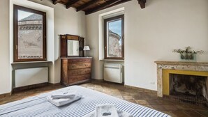 Une chambre à coucher comprenant un lit, une commode, un miroir, une cheminée et deux fenêtres donnant sur un immeuble.