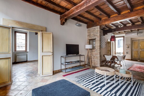 A room with a TV, a bookshelf, a dining area, and a stone wall.