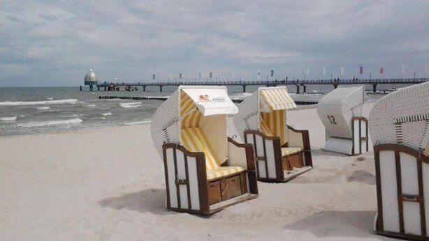 Beach chairs with striped cushions on a sandy beach, a pier with flags in the background, and a lighthouse. - Modern vacation apartment right on the beach with balcony - Residenz am Strand (Zingst)