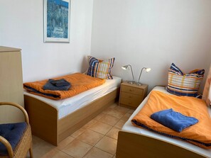 Une chambre avec deux lits simples, chacun recouvert d’une couverture orange et agrémenté d’un oreiller bleu, une table de chevet en bois avec deux lampes, une chaise en osier et un tableau encadré au mur.
