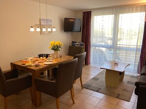 Un coin repas avec une table en bois mise pour le petit-déjeuner, un téléviseur fixé au mur et une vue sur l’extérieur grâce à de grandes fenêtres.