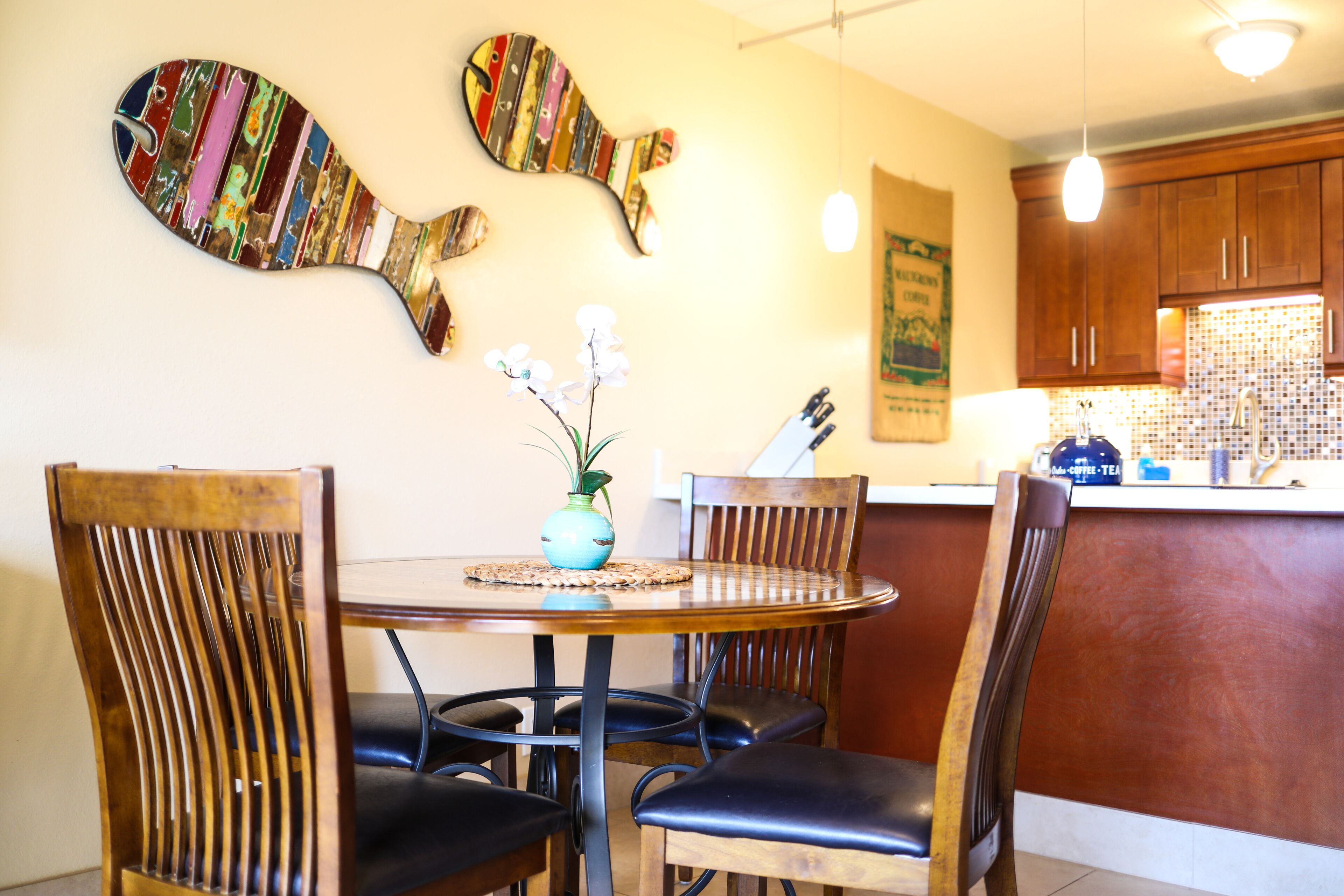 A dining area with a round wooden table, four wooden chairs, a vase with flowers, and a fish-shaped wall decoration.
