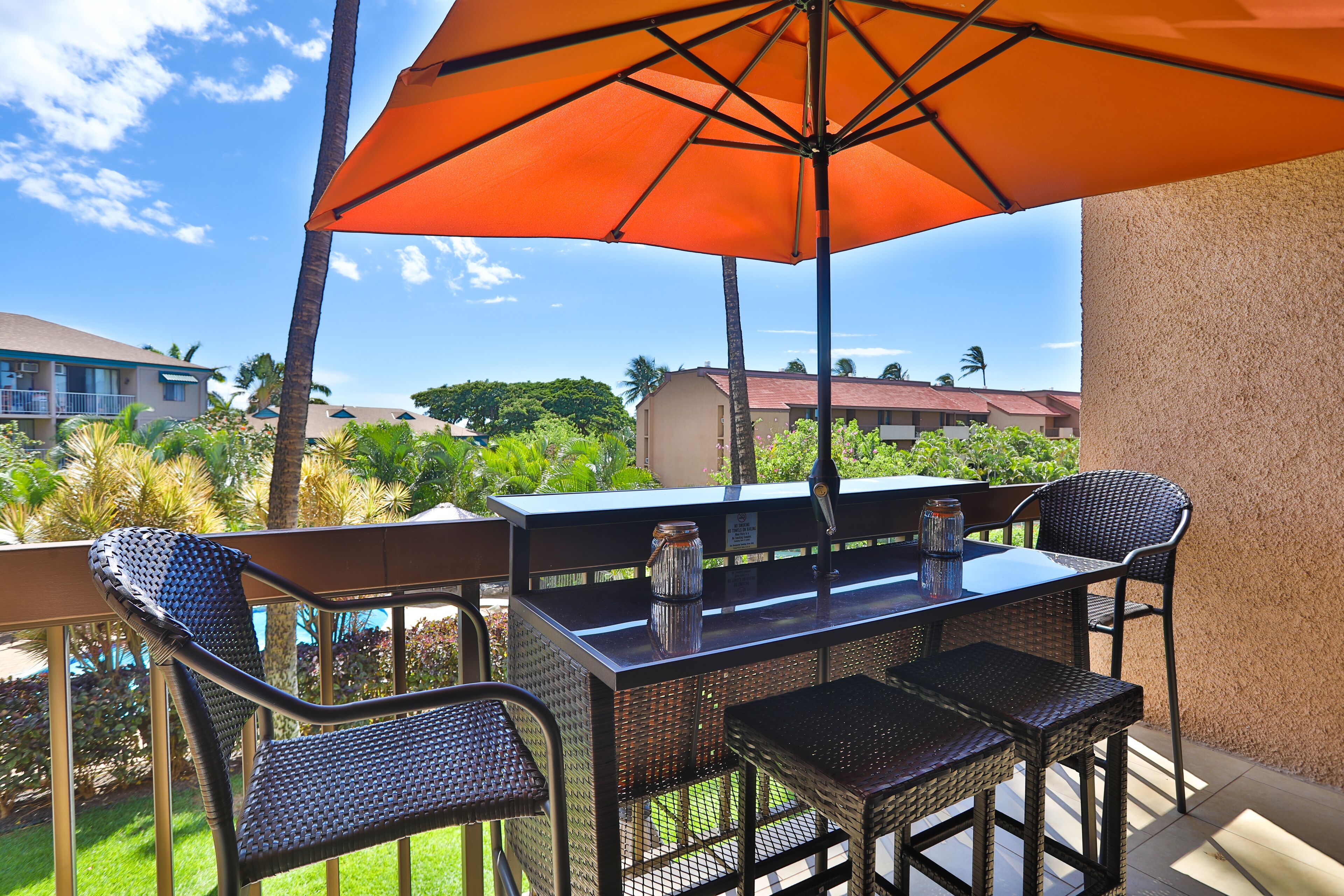 Un patio al aire libre con una mesa y sillas, un gran paraguas naranja y vistas de edificios y vegetación circundantes.