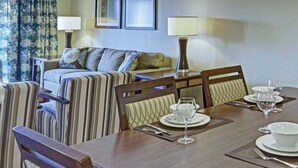 Un coin repas avec une table en bois dressée pour quatre personnes, entourée de chaises à rayures. Un canapé avec des coussins à motifs se trouve à l’arrière-plan, et deux lampes sont fixées au mur.
