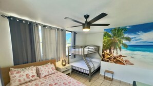 A bedroom with a bunk bed, a single bed with floral bedding, a ceiling fan, a nightstand with a lamp, and a beach-themed wall mural. - Serenity By the Beach, Private pool & El Yunque rainforest. 2BR. - 2 FULL BA. (Luquillo)