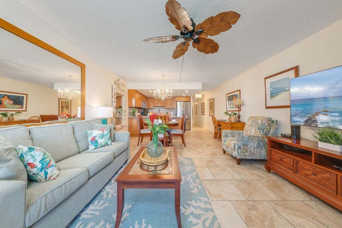 A spacious living room with a sofa, coffee table, and a television. A ceiling fan is visible, and there are framed pictures on the walls.