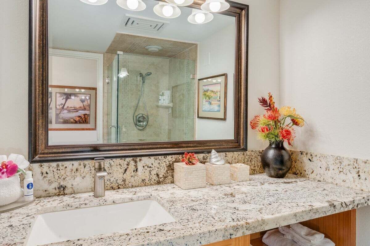 A bathroom with a large mirror, a sink, and a shower. There are framed pictures on the wall and a vase with flowers on the counter.