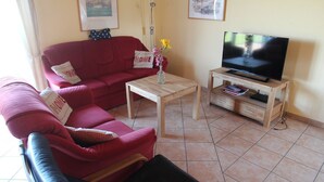 Una sala de estar con un sofá rojo, una mesa de centro de madera, un televisor sobre un mueble de madera y un florero con flores.