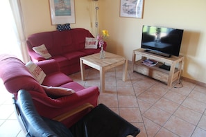 Een woonkamer met een rode bank, een houten salontafel, een tv op een houten dressoir en een vaas met bloemen.