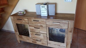 Un mueble de madera con cajones de cristal, una mesita con una taza y un mando a distancia, y un altavoz encima.