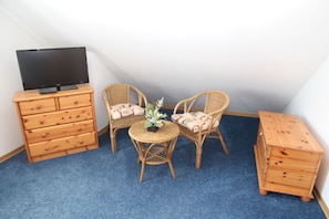 A small room with a blue carpet, two wicker chairs, a small round table with a vase, a wooden dresser, and a television on top.