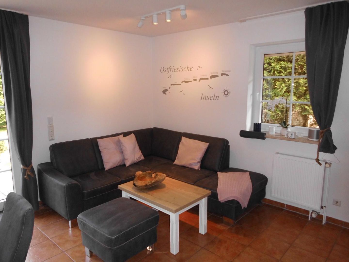 Ein gemütliches Wohnzimmer mit einer dunkelbraunen Couch, zwei Kissen, einem Holztisch und einem kleinen Hocker. Eine Wandkarte mit dem Text "Ostfriesische Inseln" ist zu sehen.