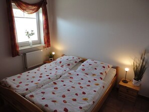 A bedroom with a double bed, a window with curtains, a radiator, a bedside table with a lamp, and a potted plant. - Friesenstern - Holiday home (100sqm) for 6 persons with garden in a quiet location of (Norden)