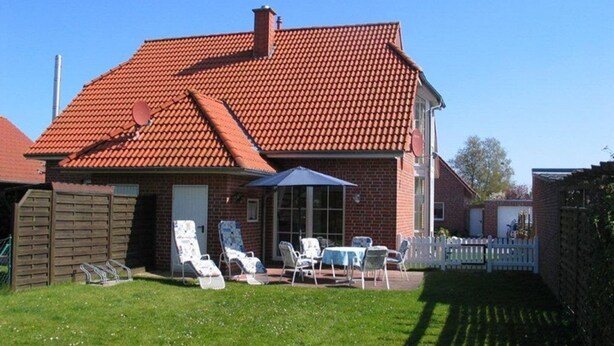 Une maison avec un toit recouvert de tuiles rouges, un patio couvert avec des meubles d’extérieur et une clôture blanche faite de piquets.