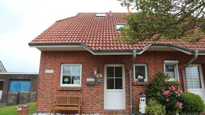Une maison en briques avec un toit recouvert de tuiles rouges, une porte d’entrée, deux fenêtres, un banc et un petit jardin agrémenté de fleurs et d’une nichoir.
