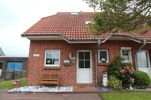 A brick house with a red-tiled roof, a front door, two windows, a bench, and a small garden with flowers and a birdhouse.