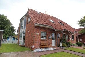 Uma casa de dois andares de tijolo, com telhado de telhas vermelhas, varanda frontal com um banco e um pequeno jardim com uma planta em vaso.