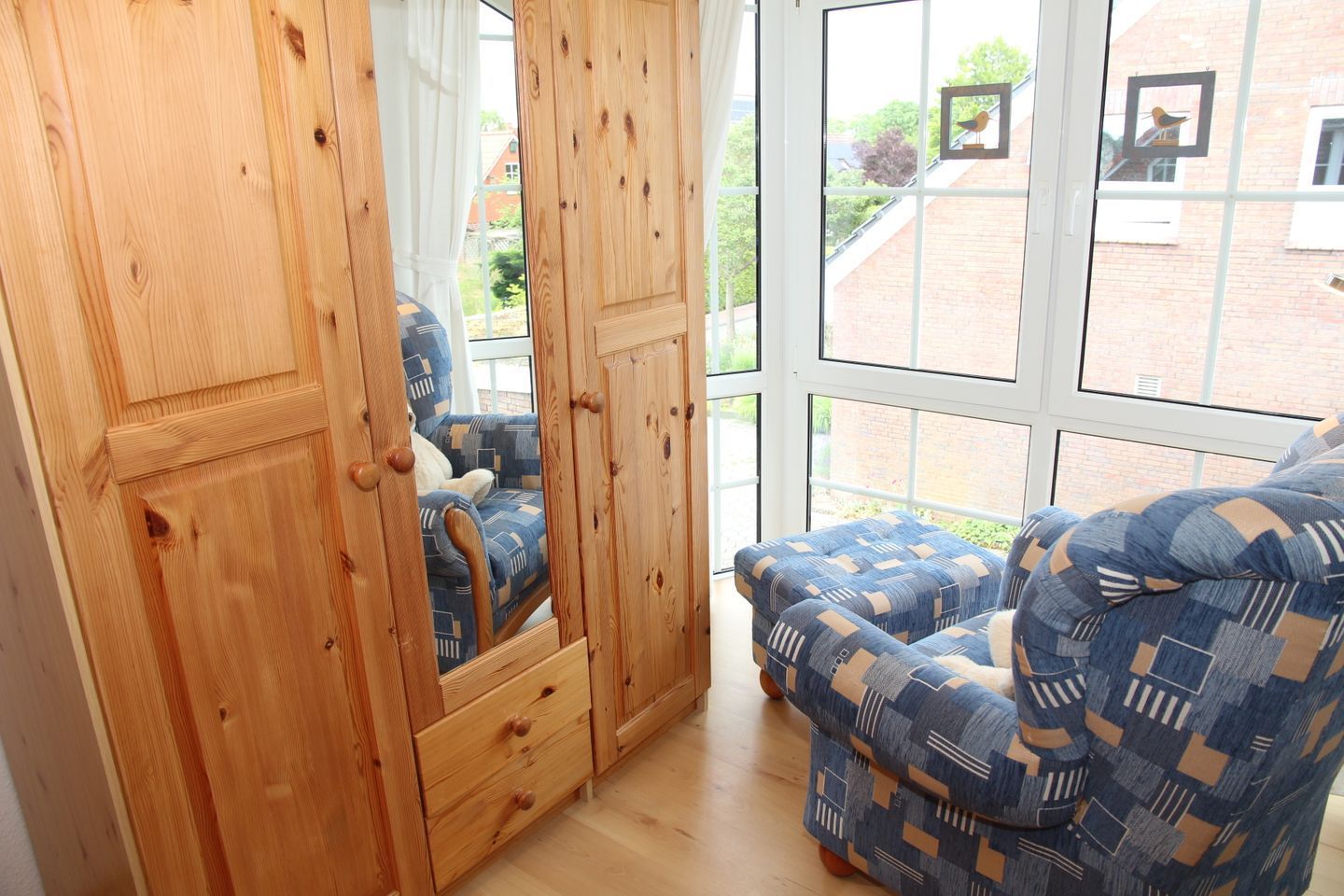 Ein Zimmer mit einem Holzschrank, einem blau gemusterten Sessel und einem großen Fenster mit Blick auf ein Backsteingebäude und Grünflächen.