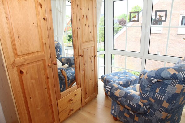 Quarto com armário de madeira, uma cadeira estofada azul com padrão e uma janela grande com vista para um edifício de tijolos e vegetação.