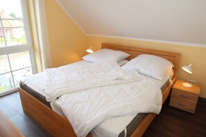 A neatly made bed in a room with a wooden headboard, a bedside table with a lamp, and a window providing a view of the outside.
