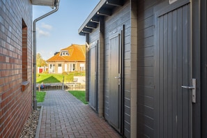 A narrow brick pathway leading to a building with grey doors, a brick wall, and a house with a red roof in the background.