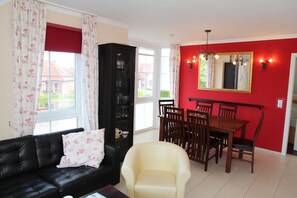 A living room with a black leather sofa, a cream-colored armchair, a dining table with wooden chairs, and a red accent wall with a mirror. - Dornröschen - Ferienwohnung mit Direktem Wasserblick (Krummhörn)