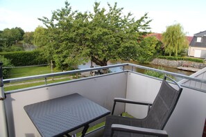 A balcony with a metal railing, a small table, and a chair. There is a view of a river and trees in the background. - Dornröschen - Ferienwohnung mit Direktem Wasserblick (Krummhörn)