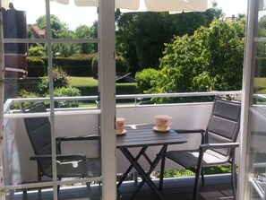A balcony with a table and chairs, overlooking a garden with trees and a hedge.