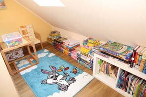 Un cuarto de juegos infantil con una alfombra azul que muestra un conejo de caricatura, un corral de madera y estantes llenos de rompecabezas y libros.