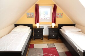 A small, triangular room with two single beds, a window with red curtains, and a radiator.