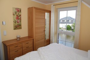 A bedroom with a bed, wooden wardrobe, and a window with a view of a house and water.