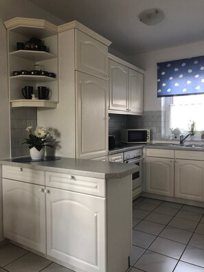 A kitchen with white cabinetry, a microwave, a stove, and a sink. There is a window with a blue curtain and a potted plant on the counter. - Flüthörn - Cozy semi-detached house with unobstructed views (Norden)