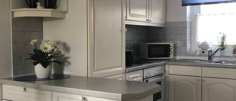 Una cocina con muebles blancos, un microondas, una estufa y un lavabo. Hay una ventana con una cortina azul y una planta en maceta sobre la encimera.