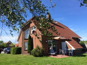 Een twee verdiepingen tellend bakstenen huis met een schuin dak, een klein voortuintje met een tafel en stoelen, en een verzorgd gazon.