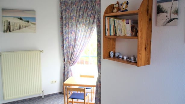 A room with a wooden chair and table, a radiator, a bookshelf with books and decorative items, and a window with curtains. - Flüthörn - Cozy semi-detached house with unobstructed views (Norden)