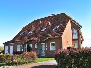 A two-story brick house with a sloped roof, a front porch, and a small garden. - Flüthörn - Cozy semi-detached house with unobstructed views (Norden)