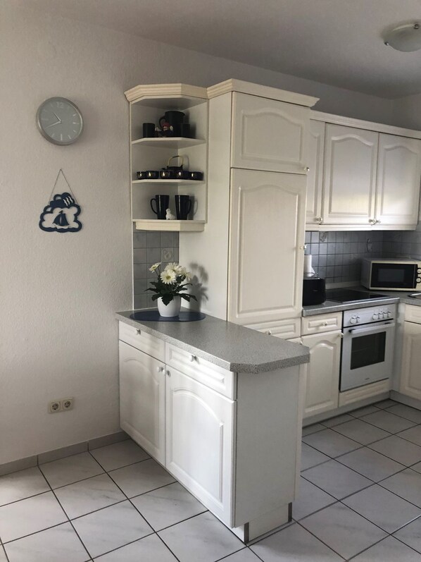 A kitchen with white cabinets, a microwave, an oven, and a clock on the wall. - Flüthörn - Cozy semi-detached house with unobstructed views (Norden)