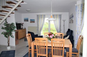 A dining area with a wooden table and chairs, a potted plant, and a view of the outdoors through large windows. - Flüthörn - Cozy semi-detached house with unobstructed views (Norden)