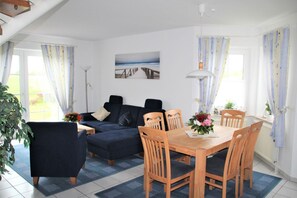 A cozy living room with a dark blue sofa, wooden dining table, and chairs. There is a large window with curtains, a floor lamp, and a painting of a pier on the wall. - Flüthörn - Cozy semi-detached house with unobstructed views (Norden)