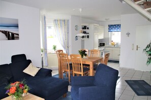 A living room with a sofa, chairs, a dining table, and a kitchen area with cabinets and a microwave. - Flüthörn - Cozy semi-detached house with unobstructed views (Norden)