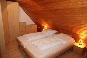 Makuuhuone, jossa on puinen, kalteva katto, valkoisilla vuodevaatteilla varustettu sänky, puinen vaatekaappi ja yöpöytä, jossa on lamppu.