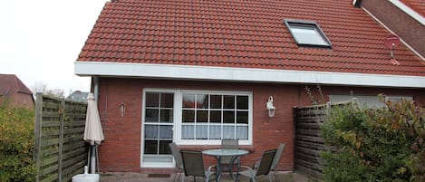 A house with a red tiled roof, a large window with a white frame, and a small skylight. There is a patio with a table and chairs, and a wooden fence in the background.