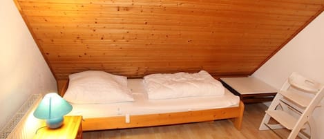 Ein kleiner, gemütlicher Raum mit Holzbalkendecke und einem Einzelbett. Es gibt einen Nachttisch mit einer Lampe und einen Stuhl.