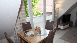 A dining area with a wooden table, brown chairs, a glass of water, and a menu. There is a TV and a lamp in the background. - Wattwunder - Schöne Ferienwohnung mit Inkludiertem Strandkorb am Strand (Norden)
