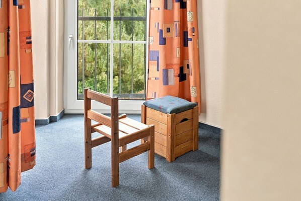 A room with a wooden chair and a small wooden stool, a window with a view of greenery, and orange patterned curtains.