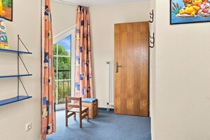 A room with a wooden door, a window with patterned curtains, a blue carpet, a wooden chair, and a shelf with a picture. - Sandrose - Semi-detached house (85 m²) for 4 people with a garden right on the beach (Norden)