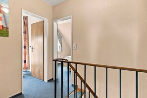 A hallway with a wooden door leading to a room, a staircase with a wooden handrail, and a wall-mounted light switch.