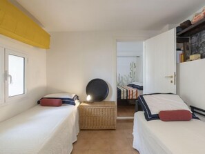 Une petite pièce bien éclairée, avec deux lits simples, une commode en osier et une porte donnant sur une autre pièce.