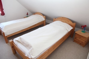 Deux lits simples en bois, recouverts de draps blancs, se trouvent dans une pièce dont le plafond est en pente. Une petite table de chevet en bois, sur laquelle est posée une lampe, est également présente.